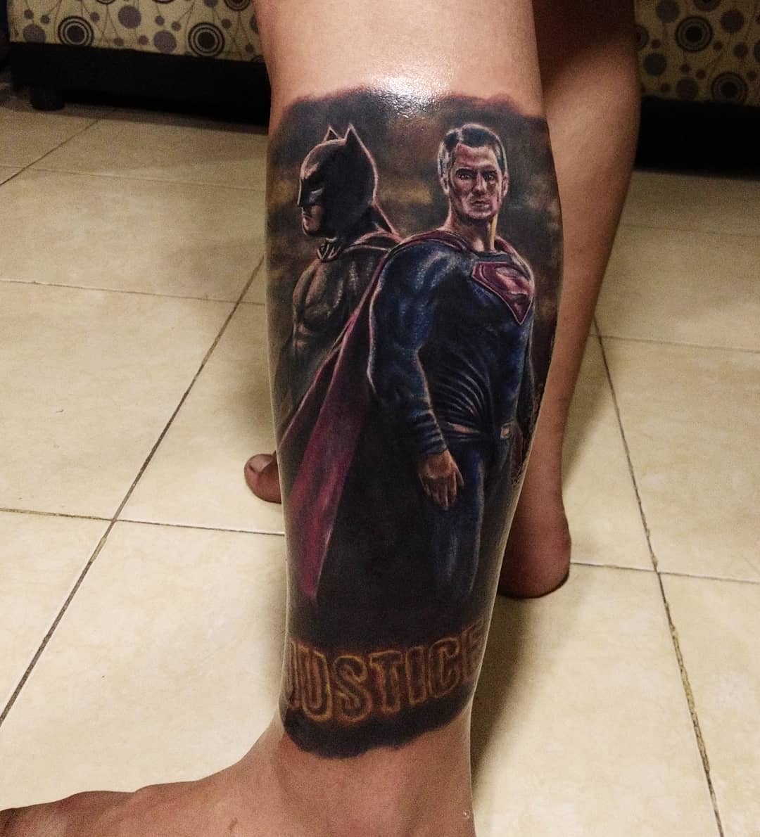 Tattoo of Batman and Superman on a calf with the word "Justice" at the bottom